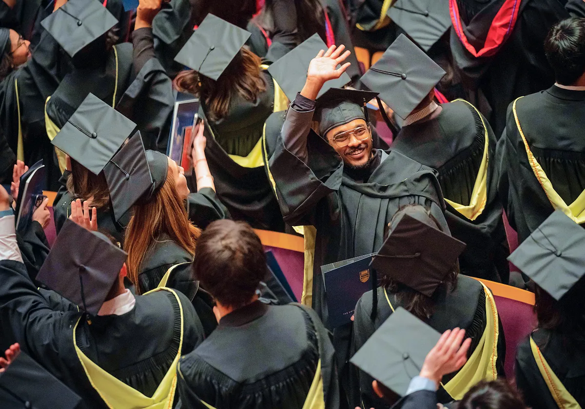 A graduating student wearing a cap and gown smiling and waving in a crowd of fellow graduates.
