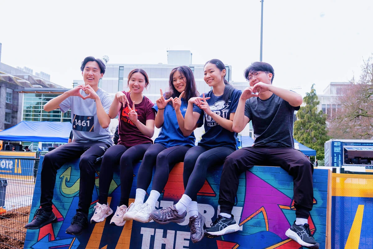 Five Storm the Wall student competitors sitting on the wall, each making a different hand gesture.
